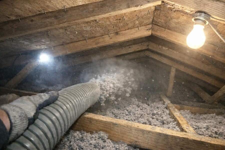 A blown-in insulation hose filling a joist bay with loose gray insulation in a low attic